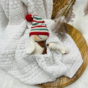 May include: A white knitted baby blanket with a decorative pattern, featuring a crocheted duck wearing a red, green, and white striped hat. A small crocheted duck foot is also visible. The items are arranged on a wooden tray.