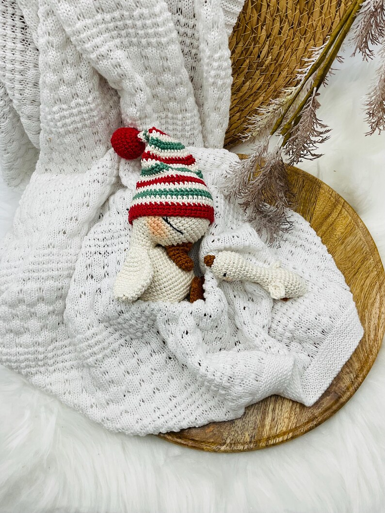 May include: A white knitted baby blanket with a decorative pattern, featuring a crocheted duck wearing a red, green, and white striped hat. A small crocheted duck foot is also visible. The items are arranged on a wooden tray.