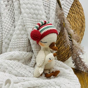 May include: A handmade crochet goose doll wearing a red, green, and white striped hat with a red pom-pom. The goose is white with brown feet and a small baby goose attached. The doll is set against a white knitted blanket.