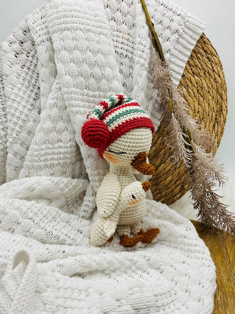 May include: A handmade crochet goose doll wearing a red, green, and white striped hat with a red pom-pom. The goose is white with brown feet and a small baby goose attached. The doll is set against a white knitted blanket.