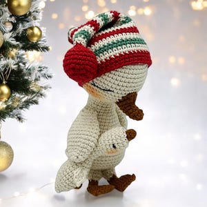 May include: A crocheted goose doll wearing a striped red, green, and white hat with ear muffs. The goose is holding a smaller goose. The doll is cream-colored with brown accents and is next to a Christmas tree with gold ornaments.