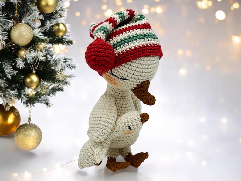 May include: A crocheted goose doll wearing a striped red, green, and white hat with ear muffs. The goose is holding a smaller goose. The doll is cream-colored with brown accents and is next to a Christmas tree with gold ornaments.