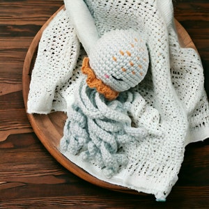 Op de afbeelding: Een gehaakt octopus speelgoed met een lichtblauw lichaam en oranje accenten. De octopus ligt op een wit gehaakt deken.