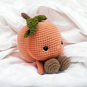 May include: A crocheted peach plush toy with a green leaf on top and two brown crocheted feet. The peach has a stitched-on face with black eyes.