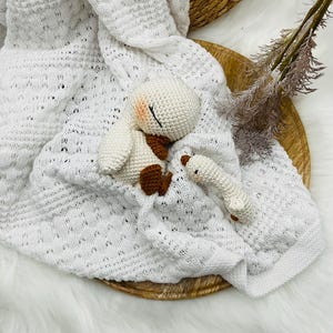 May include: A white knitted baby blanket with a textured pattern, paired with a crocheted bunny and a small rattle. The bunny has brown accents and closed eyes. The items are arranged on a wooden tray.