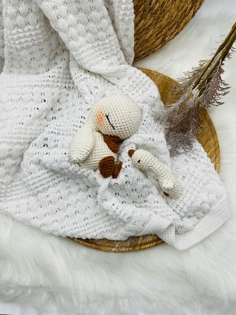 May include: A white knitted baby blanket with a textured pattern, paired with a crocheted bunny and a small rattle. The bunny has brown accents and closed eyes. The items are arranged on a wooden tray.