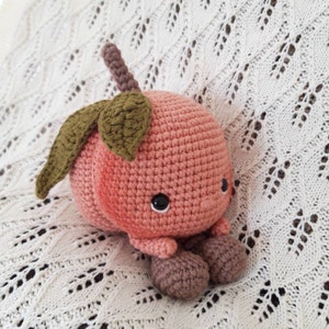 May include: A crocheted peach plush toy with a green leaf stem and brown legs. The peach has a pink and white color gradient and black eyes.