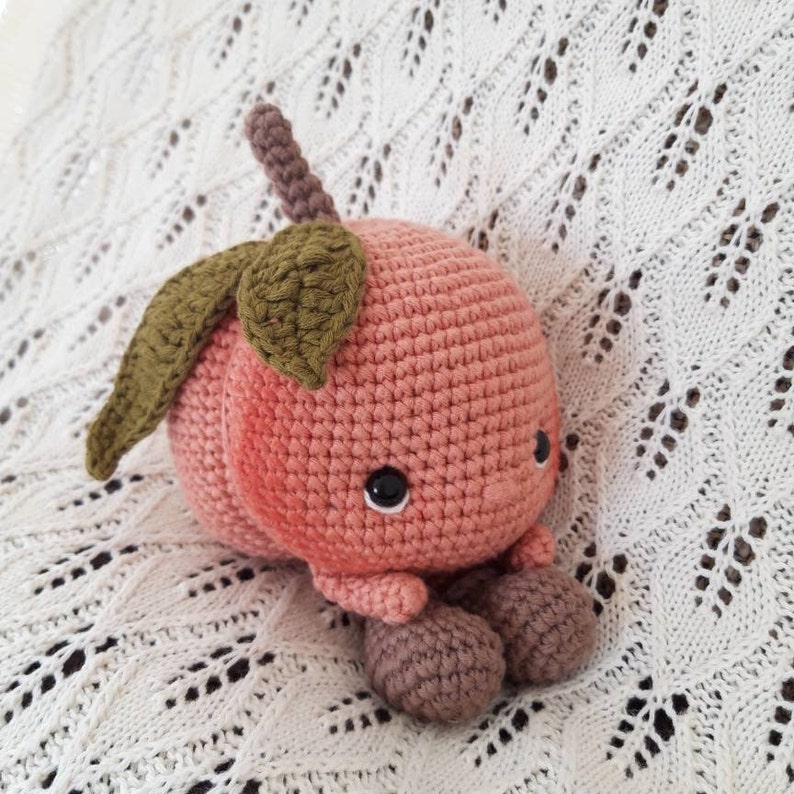 May include: A crocheted peach plush toy with a green leaf stem and brown legs. The peach has a pink and white color gradient and black eyes.