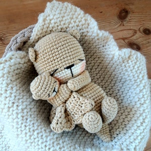 May include: A crocheted teddy bear nestled in a cream-coloured knitted blanket. The bear is light brown with closed eyes and a small pink blush on its cheeks. The blanket has a scalloped edge and is in a woven basket.