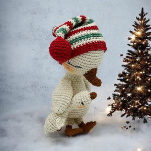 May include: A handmade crochet goose wearing a striped red, green, and white hat with a red pom-pom. The goose is holding a smaller version of itself. The goose has brown feet and a brown beak. A Christmas tree with lights is in the background.