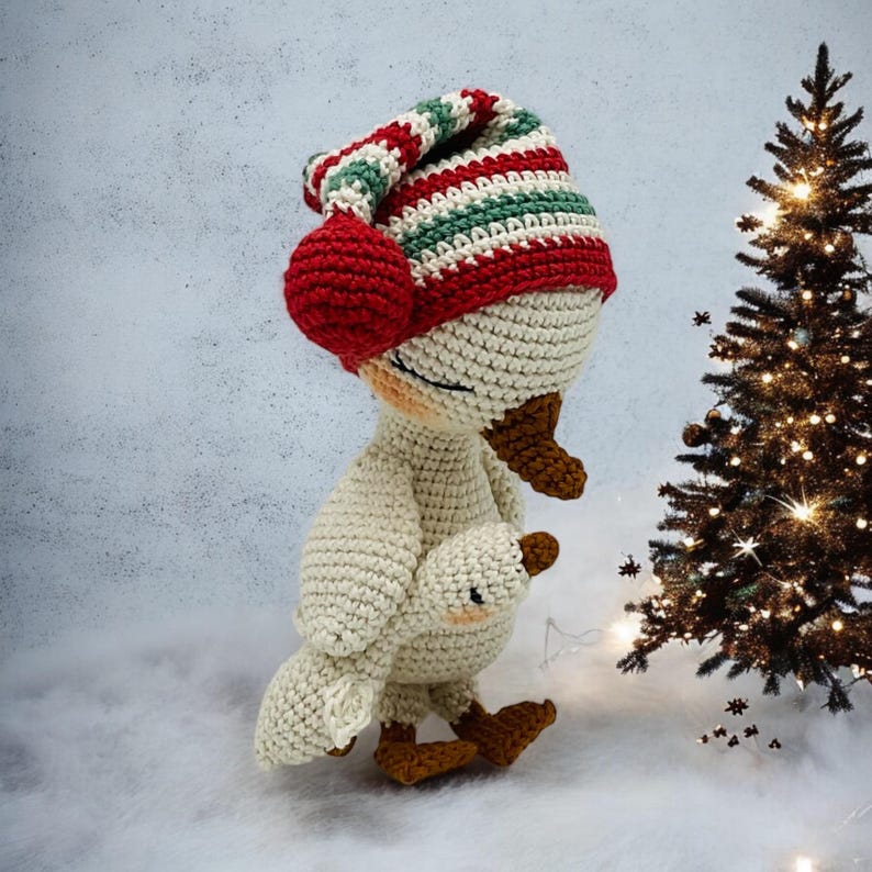 May include: A handmade crochet goose wearing a striped red, green, and white hat with a red pom-pom. The goose is holding a smaller version of itself. The goose has brown feet and a brown beak. A Christmas tree with lights is in the background.
