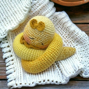 Può includere: Un giocattolo amigurumi all'uncinetto giallo e arancione a forma di serpente arrotolato con un fiocco giallo sulla testa. Il giocattolo è sdraiato su una coperta di uncinetto bianca.