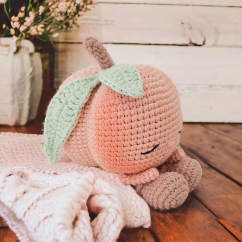 May include: A crocheted peach-colored plush toy with a green leaf on top. The toy is lying on a white crocheted blanket.