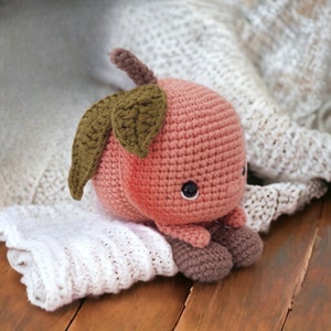 May include: A crocheted plush toy shaped like a cherry with a green stem and leaves. The cherry is pink and has a cute face with black eyes. The toy is sitting on a white knitted blanket.