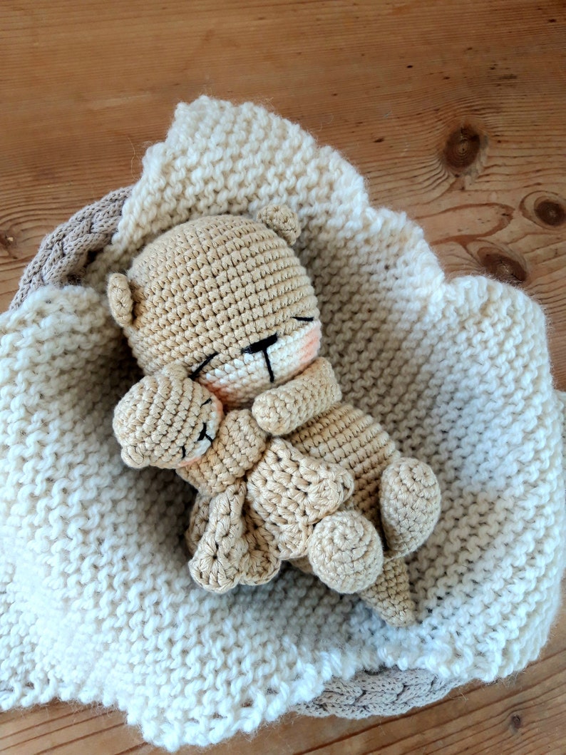 May include: A crocheted teddy bear nestled in a cream-coloured knitted blanket. The bear is light brown with closed eyes and a small pink blush on its cheeks. The blanket has a scalloped edge and is in a woven basket.
