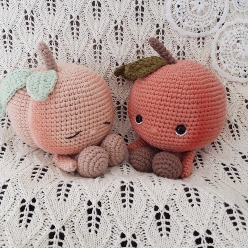 May include: Two crocheted peach-colored plush toys shaped like peaches with embroidered eyes and smiling mouths. The toys are sitting on a white crocheted blanket.