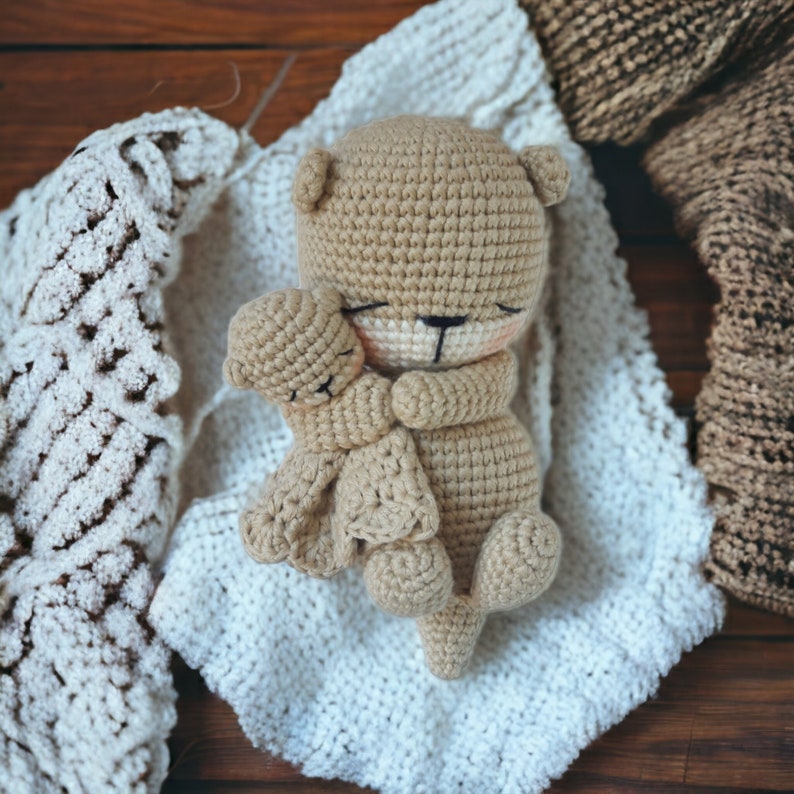 May include: A light brown crocheted teddy bear embracing a smaller teddy bear, resting on a white crocheted blanket. The image is a close-up, with a warm, inviting feel. The texture of the crochet is clearly visible.