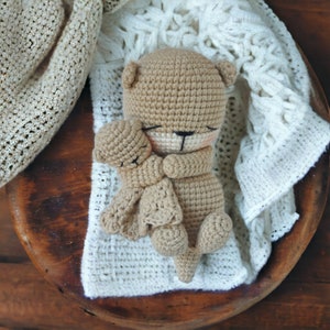 May include: A light brown crocheted teddy bear embracing a smaller bear. The bears rest on a white, textured blanket, within a wooden bowl. The bear has a black nose and closed eyes, suggesting sleep.