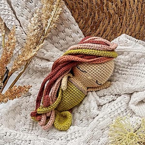 May include: A handmade crocheted mermaid doll with a beige head, green tail, and long, colourful hair in shades of red, pink, and yellow. The doll is lying on a white, textured blanket. A unique, handcrafted item.