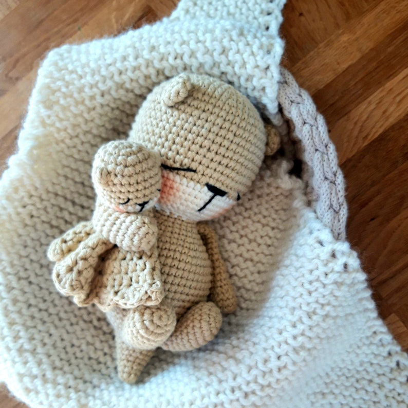 May include: A light brown crocheted teddy bear with closed eyes and rosy cheeks, cuddling a smaller version of itself. The bear rests in a cream-coloured, knitted blanket. The bear has black stitched details and a small, attached scarf.