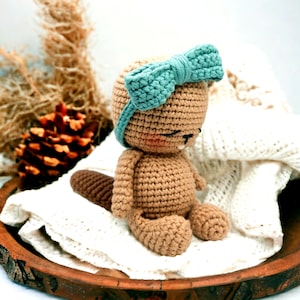 May include: A crocheted beaver toy wearing a teal blue bow. The beaver is sitting on a white crocheted blanket on a wooden surface.