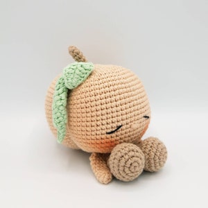 May include: A crocheted peach plush toy with a green leaf on top. The peach has a closed-eye face and two small crocheted arms.