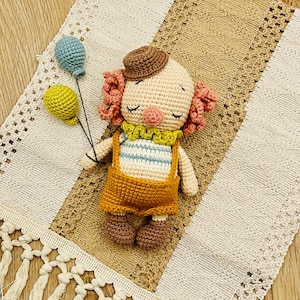 May include: A crocheted clown doll with pink hair, a brown hat, and a yellow and blue striped shirt. The doll is holding two balloons, one yellow and one blue. The doll is lying on a crocheted blanket with fringe.