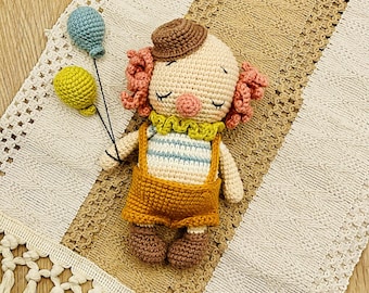 Puede incluir: Una muñeca payaso de crochet con pelo rosa, un sombrero marrón y una camisa a rayas amarilla y azul. La muñeca sostiene dos globos, uno amarillo y otro azul. La muñeca está tumbada sobre una manta de crochet con flecos.