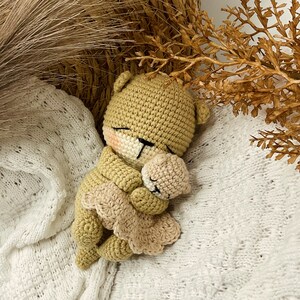 May include: A crocheted teddy bear doll in a sleeping pose, with closed eyes and a light pink blush on its cheeks. The bear is light brown, wearing a light tan dress. It is holding a smaller bear. The doll is lying on a white, textured blanket.