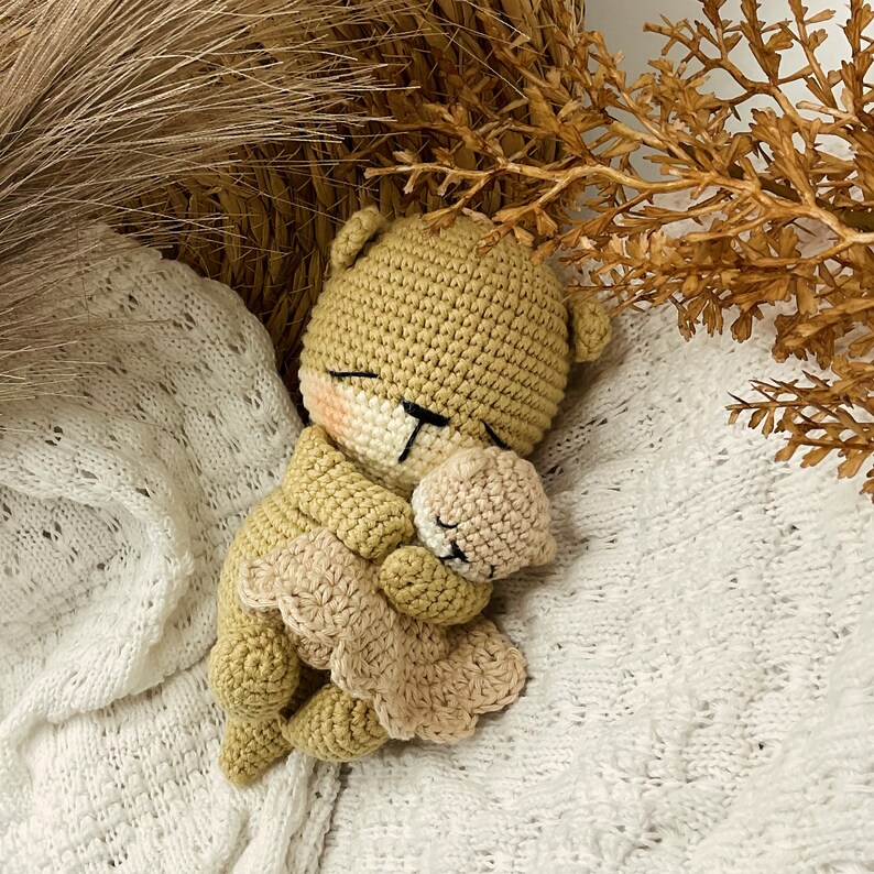May include: A crocheted teddy bear doll in a sleeping pose, with closed eyes and a light pink blush on its cheeks. The bear is light brown, wearing a light tan dress. It is holding a smaller bear. The doll is lying on a white, textured blanket.