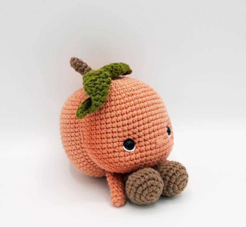 May include: A crocheted peach plush toy with a green leaf on top and brown crocheted feet. The peach has a cute face with black eyes.