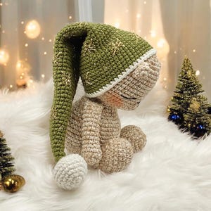 May include: A crocheted teddy bear wearing a green and gold crocheted Santa hat with a white pom-pom. The bear is sitting on a white fluffy surface.