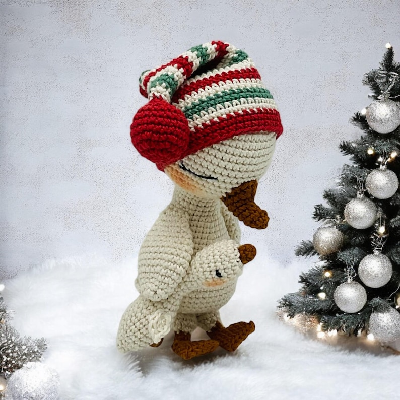 May include: A handmade crochet goose wearing a striped red, green, and white hat with a red pom-pom. The goose is holding a smaller version of itself. The background includes a Christmas tree with silver ornaments.