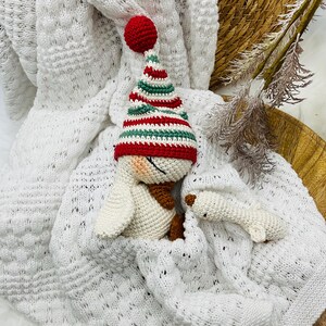 May include: A crocheted snowman wearing a red, green, and white striped hat and a brown scarf, nestled in a white knitted blanket. A small, white crocheted snowman is also present. The scene is set on a wooden tray.