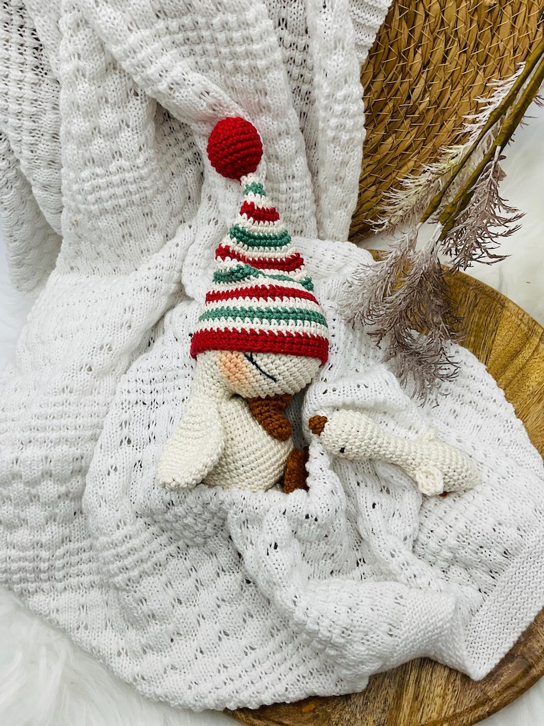 May include: A crocheted snowman wearing a red, green, and white striped hat and a brown scarf, nestled in a white knitted blanket. A small, white crocheted snowman is also present. The scene is set on a wooden tray.