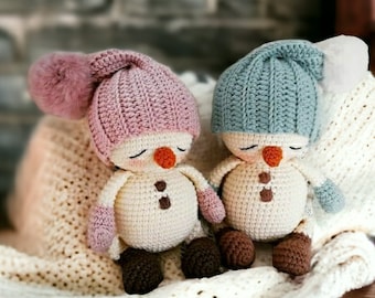 Puede incluir: Dos adornos de muñeco de nieve de crochet con caras dormidas. Un muñeco de nieve lleva un gorro rosa con un pompón blanco y el otro lleva un gorro azul con un pompón blanco.