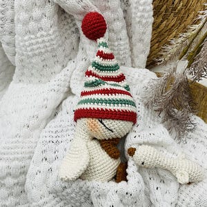 May include: A handmade crochet snowman doll with a red, green, and white striped hat and a matching red pom-pom. The snowman has a brown scarf and is lying on a white knitted blanket. A smaller snowman is next to it.