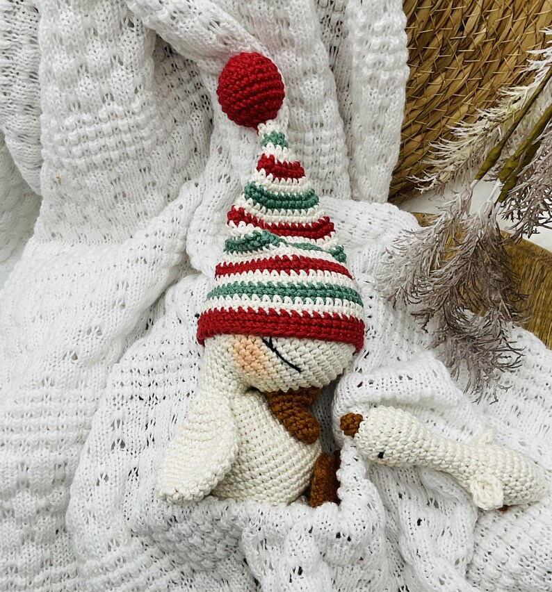 May include: A handmade crochet snowman doll with a red, green, and white striped hat and a matching red pom-pom. The snowman has a brown scarf and is lying on a white knitted blanket. A smaller snowman is next to it.