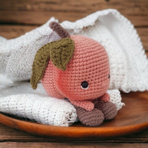 May include: A crocheted amigurumi toy shaped like a cherry with a green leaf. The cherry is pink and has a brown stem. The toy is sitting on a white crocheted blanket.
