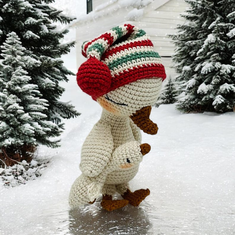 May include: A crocheted goose wearing a striped red, green, and white hat with a red pom-pom. The goose is holding a smaller crocheted goose. The background shows a snowy winter scene with evergreen trees.