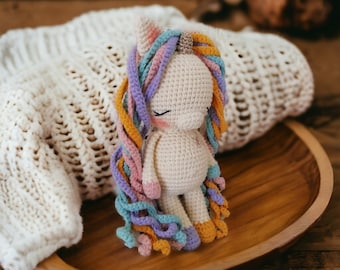 May include: A crocheted unicorn toy with a white body, purple and pink yarn mane and tail, and a yellow horn. The unicorn is sitting on a wooden surface.