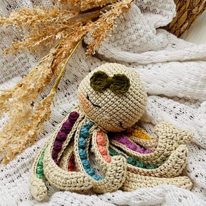 May include: A crocheted octopus toy in beige with colorful tentacle accents. The octopus has a smiling face and a small green bow. The tentacles feature rainbow-colored details. The toy is handmade and sits on a white, textured surface.