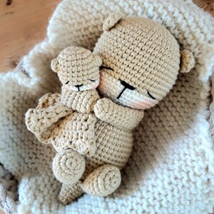 May include: A pair of crocheted teddy bears, one large and one small, are nestled together on a cream blanket. The bears are light brown with black eyes and pink accents. The blanket has a textured, woven appearance.