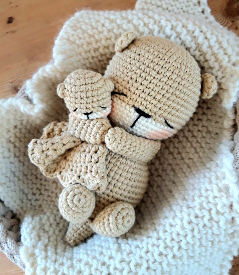 May include: A pair of crocheted teddy bears, one large and one small, are nestled together on a cream blanket. The bears are light brown with black eyes and pink accents. The blanket has a textured, woven appearance.