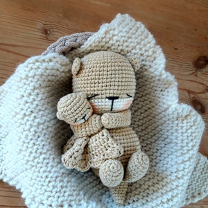 May include: A crocheted teddy bear in a cream-coloured blanket. The bear is light brown with black eyes and a pink nose. The blanket is a thick knit with a light brown border. The bear is nestled in the blanket.
