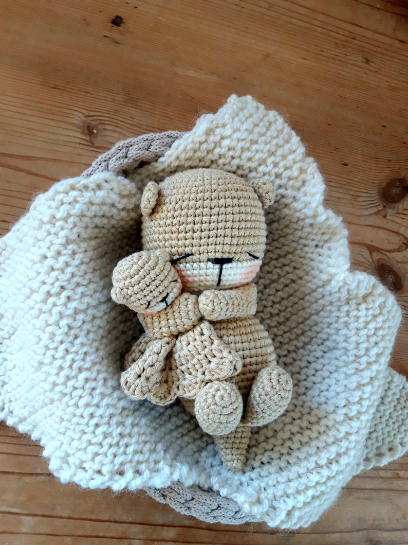 May include: A crocheted teddy bear in a cream-coloured blanket. The bear is light brown with black eyes and a pink nose. The blanket is a thick knit with a light brown border. The bear is nestled in the blanket.