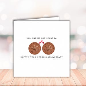 May include: A square white greeting card featuring two copper-colored British two pence coins side by side. Above the coins, the text reads YOU AND ME ARE MEANT 2p with three small red hearts. Below the coins, the text reads HAPPY 7 YEAR WEDDING ANNIVERSARY.