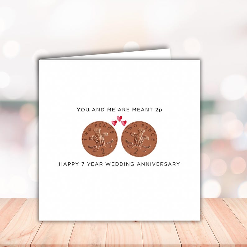 May include: A square white greeting card featuring two copper-colored British two pence coins side by side. Above the coins, the text reads YOU AND ME ARE MEANT 2p with three small red hearts. Below the coins, the text reads HAPPY 7 YEAR WEDDING ANNIVERSARY.