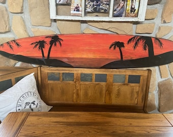 May include: A wooden surfboard painted with a sunset scene and palm trees. The surfboard is hanging on a stone wall.