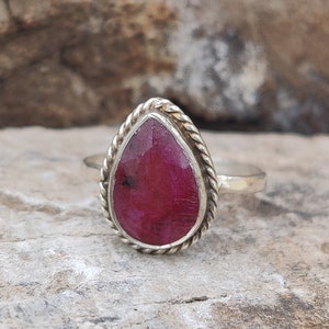 Puede incluir: Un anillo de plata con una piedra preciosa de rubí en forma de lágrima. El anillo tiene una banda torcida y un engaste de bisel.